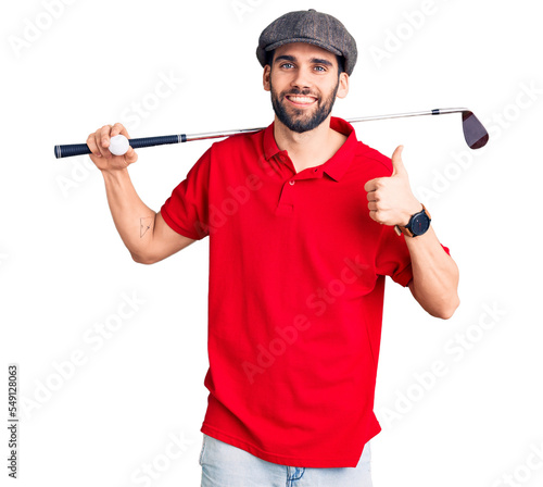 Young handsome man with beard playing golf holding club and ball smiling happy and positive, thumb up doing excellent and approval sign
