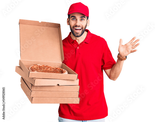 Young handsome man with beard wearing delivery uniform holding boxes with pizza celebrating victory with happy smile and winner expression with raised hands photo