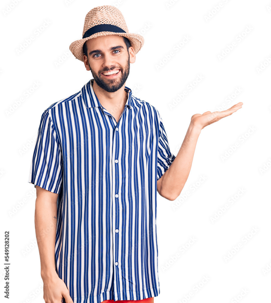 Young handsome man with beard wearing summer hat and striped shirt smiling cheerful presenting and pointing with palm of hand looking at the camera. Young handsome man with beard wearing summer hat and striped shirt smiling cheerful presenting and pointing with palm of hand looking at the camera.