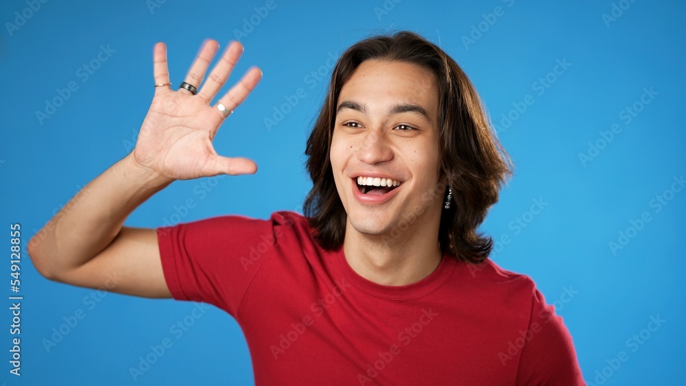 Portrait of happy smiling Hispanic gender fluid young man 20s smiling and waving meet greet with hand as notices someone isolated on blue background. Portrait of happy smiling Hispanic gender fluid young man 20s smiling and waving meet greet with hand as notices someone isolated on blue background.