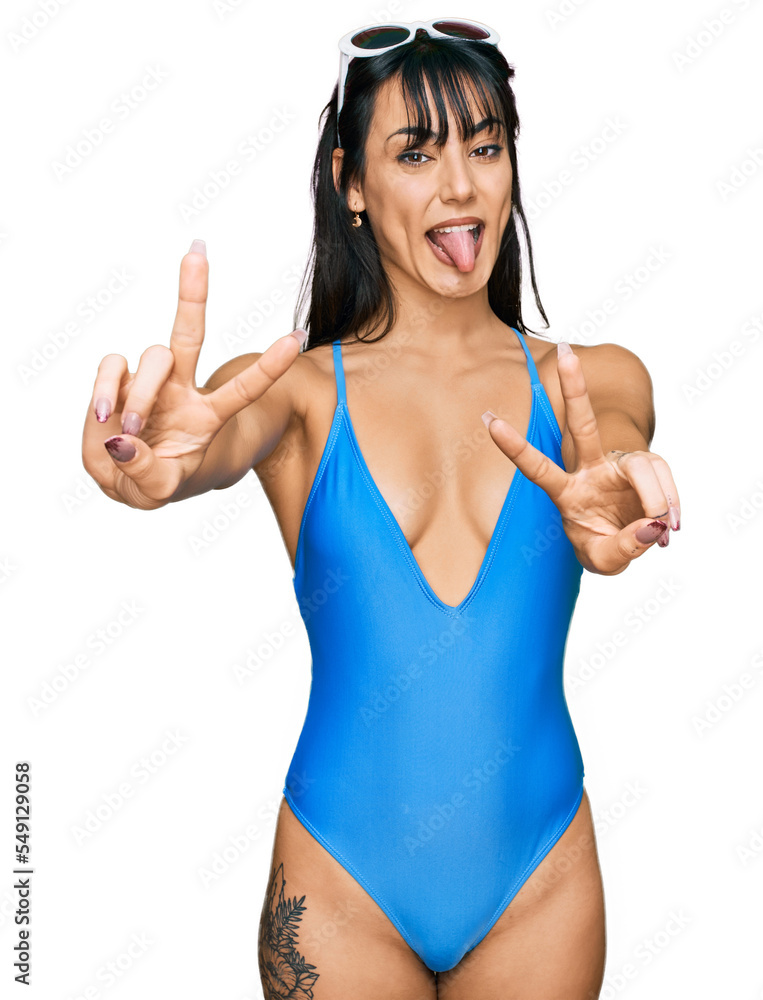 Young hispanic woman wearing swimsuit and sunglasses smiling with tongue out showing fingers of both hands doing victory sign. number two. Young hispanic woman wearing swimsuit and sunglasses smiling with tongue out showing fingers of both hands doing victory sign. number two.