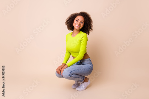 Full body photo of cute young lady curly hair squat crouch beaming smile dressed stylish yellow clothes isolated on beige color background