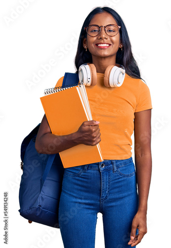 Young indian girl holding student backpack and books looking positive and happy standing and smiling with a confident smile showing teeth
