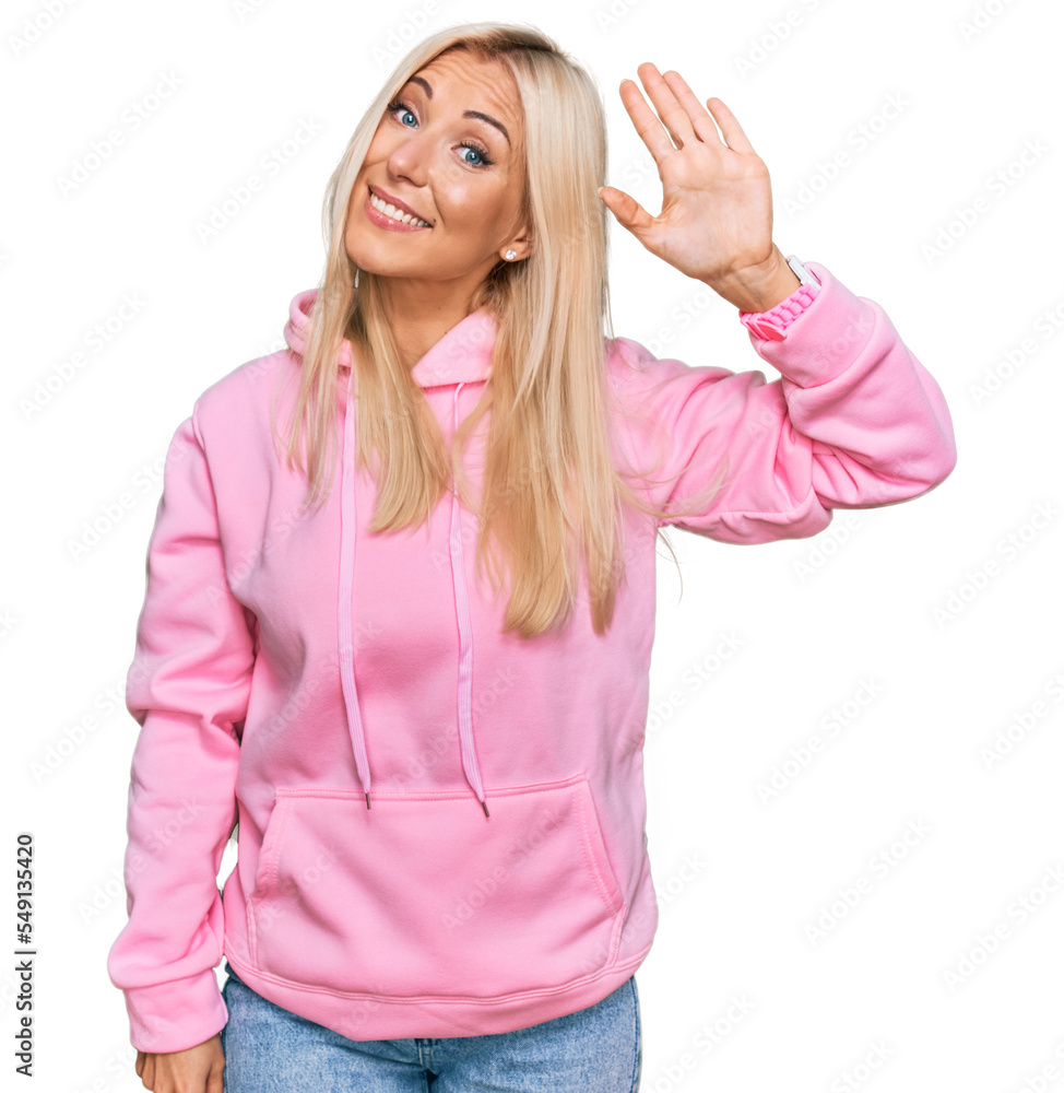 Young blonde woman wearing casual sweatshirt waiving saying hello happy and smiling, friendly welcome gesture Young blonde woman wearing casual sweatshirt waiving saying hello happy and smiling, friendly welcome gesture