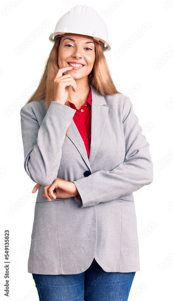 Beautiful young woman wearing architect hardhat looking confident at the camera with smile with crossed arms and hand raised on chin. thinking positive. Beautiful young woman wearing architect hardhat looking confident at the camera with smile with crossed arms and hand raised on chin. thinking positive.