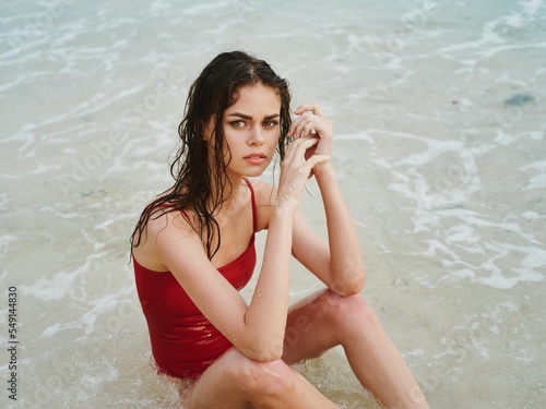 Obraz na plátně A woman with wet hair sits on the sand and looks at the camera, a slender tanned