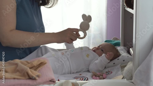 Young woman and her cute baby daughter playing with rattle toy on changing table photo