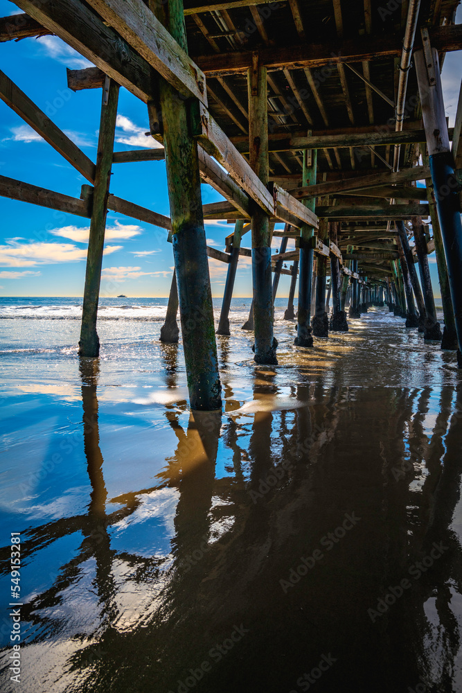 Wet pier pilings under San Clement Boardwalk at low tide with reflections on the shiny wet beach surface, abstract geometry of the crisscrossing poles against the blue cloudy sky in the background