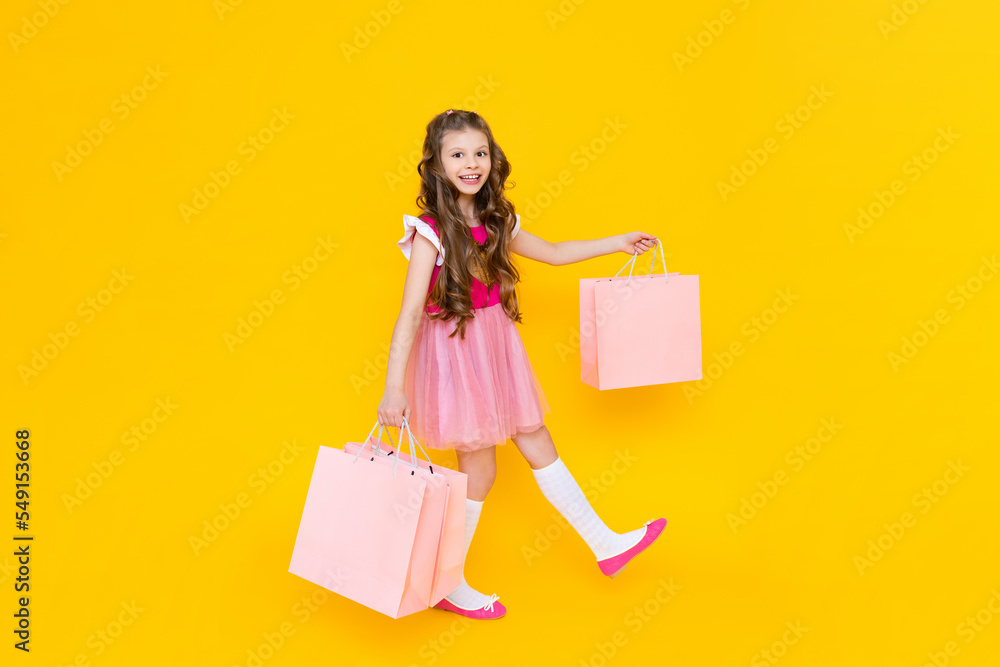 A child holds paper bags with purchases after shopping. Sale and shopping for little girls. A charming girl holds gift bags on a yellow isolated background. A child holds paper bags with purchases after shopping. Sale and shopping for little girls. A charming girl holds gift bags on a yellow isolated background.