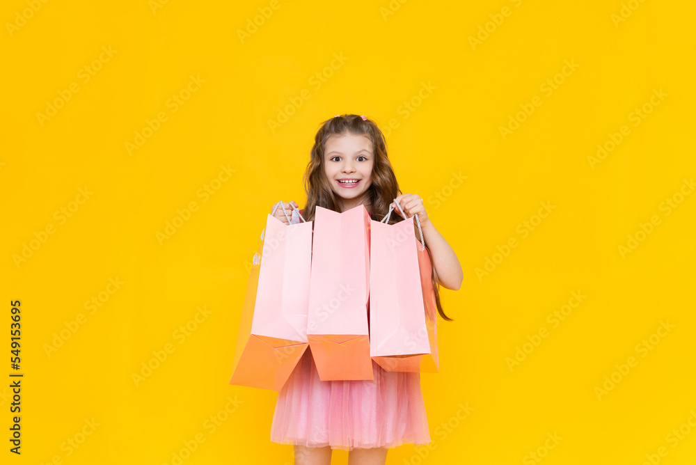 A child holds paper bags with purchases after shopping. Sale and shopping for little girls. A charming girl holds gift bags on a yellow isolated background. A child holds paper bags with purchases after shopping. Sale and shopping for little girls. A charming girl holds gift bags on a yellow isolated background.