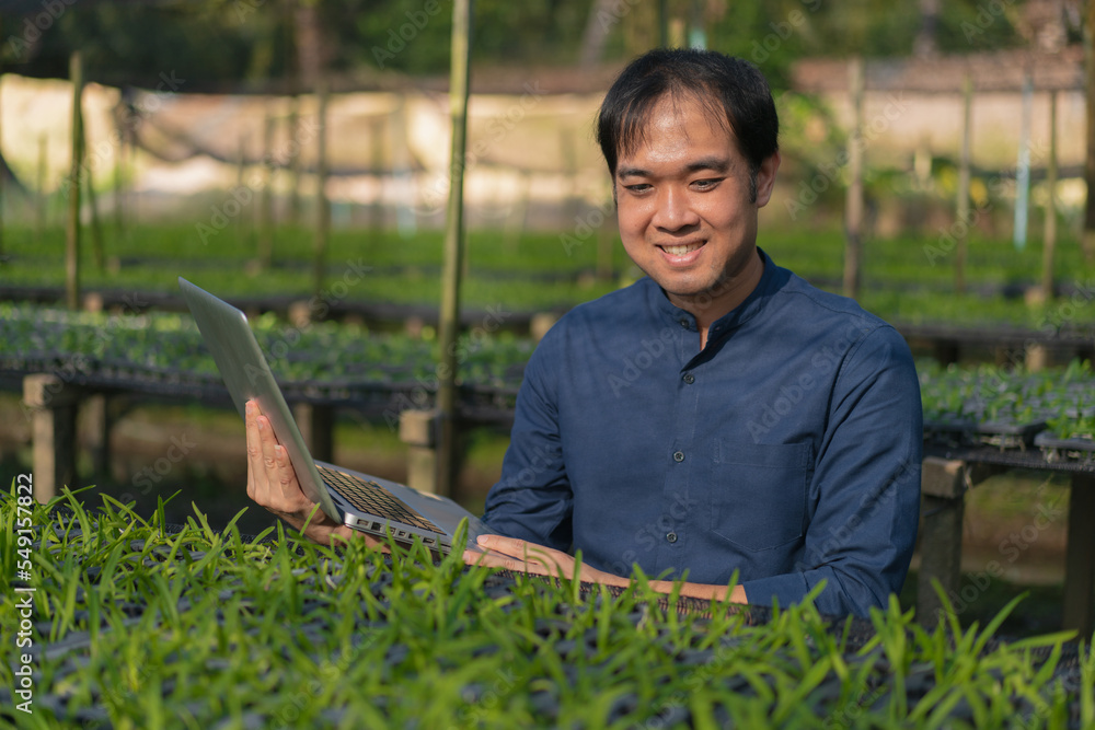 Young man farmer working at his orchid farm. Young man notes the information on the notepad to track the growth of orchid. Portrait asian small business owner of orchid gardening farm. Young man farmer working at his orchid farm. Young man notes the information on the notepad to track the growth of orchid. Portrait asian small business owner of orchid gardening farm.