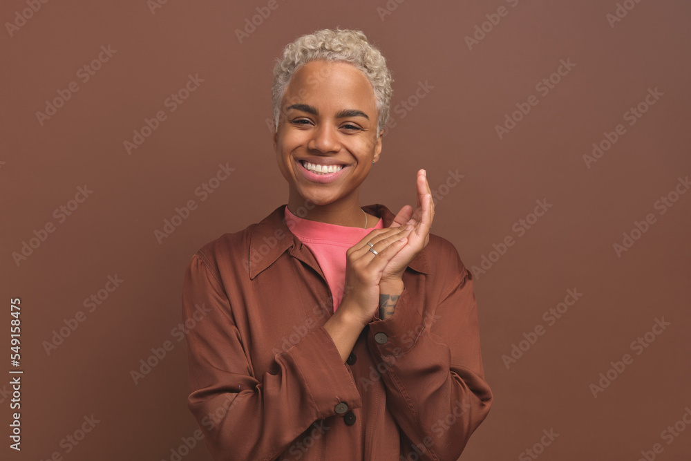 Young charismatic contented African American woman zoomer generation dressed in casual style claps hands and smiles cutely looks at camera stands on brown studio background. happiness, optimism, joy Young charismatic contented African American woman zoomer generation dressed in casual style claps hands and smiles cutely looks at camera stands on brown studio background. happiness, optimism, joy