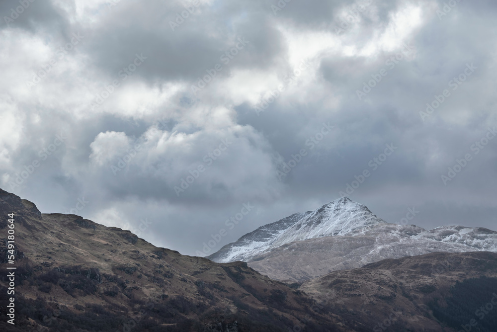 Landscape image of snowcapped Ben Lomond mountain peak in Scottish Highlands with dramatic moody sky above