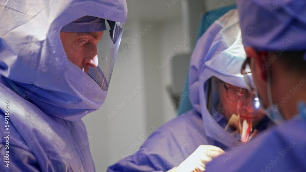 Focused doctors looking down at the patient on operational table. Close up portraits of surgeons wearing the protective suits. Focused doctors looking down at the patient on operational table. Close up portraits of surgeons wearing the protective suits.