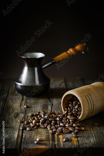 A wooden jar of coffee beans is lying on its side. Coffee beans scattered on the wooden tabletop. A copper turkish coffee pot with a long wooden handle in the background.  photo