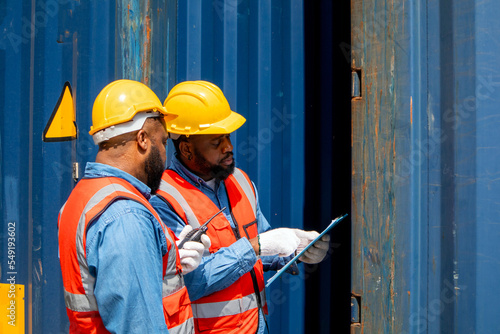 Two attractive black workers in safety helmets and uniforms work at the container port terminal. Two male engineers are checking the products in the cargo container before being exported to the port.