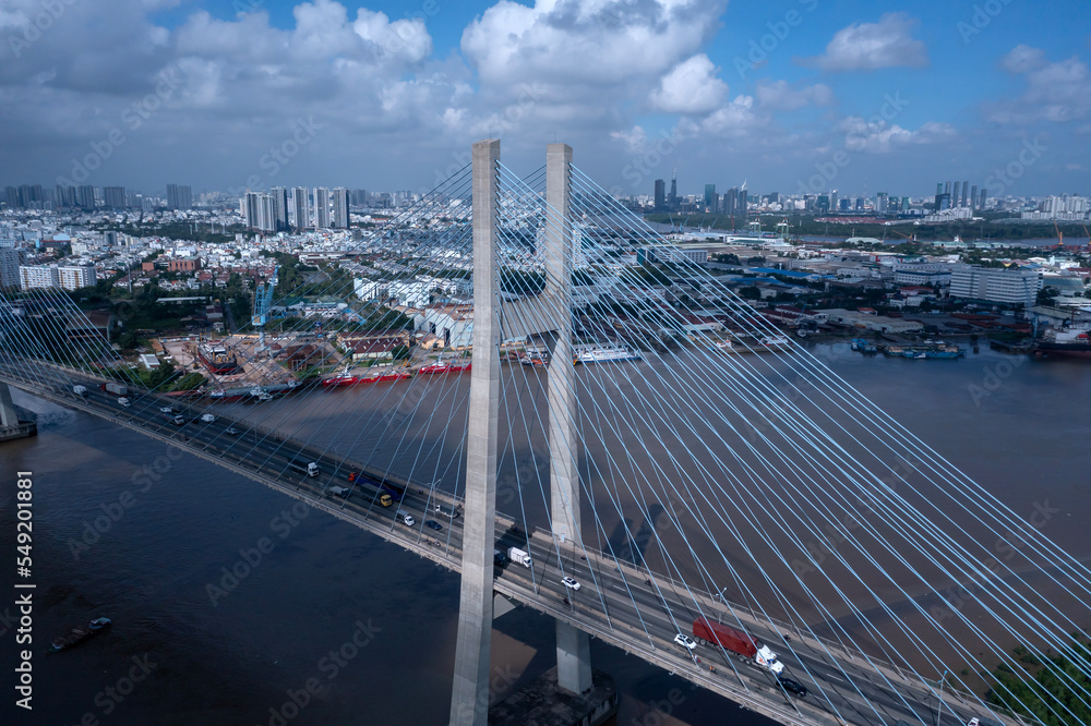 Aerial view of Phu My Bridge over Saigon river with road and river transportation on a sunny day. Cables catch light. High angle featuring roadway and heavy vehicles and Ho Chi Minh City skyline 