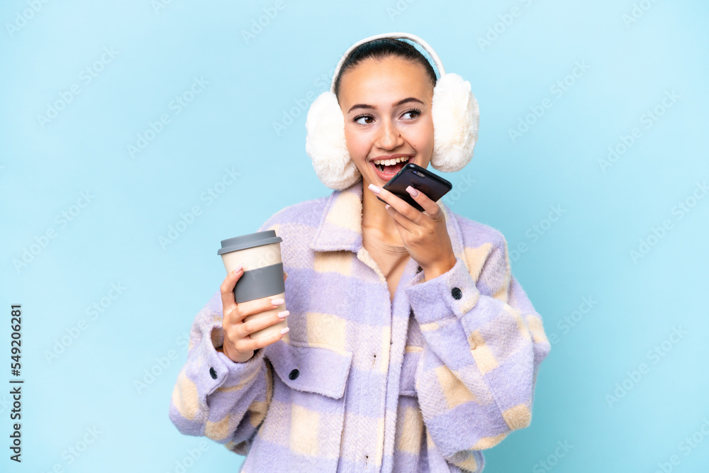 Young Arab woman wearing winter muffs isolated on blue background holding coffee to take away and a mobile Young Arab woman wearing winter muffs isolated on blue background holding coffee to take away and a mobile