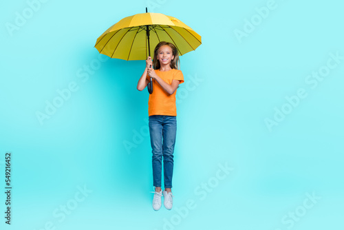 Full body length photo of little pupil girl wear stylish outfit rainy weather fly air trampoline hold umbrella isolated on aquamarine color background