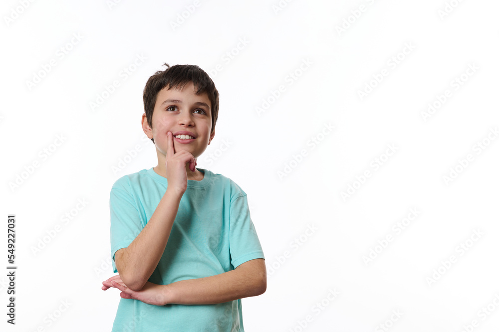 Happy teenage boy wearing turquoise t-shirt, holding his hand at chin and cutely smiling, thoughtfully looking at aside, isolated over white background with copy ad space for promotional text Happy teenage boy wearing turquoise t-shirt, holding his hand at chin and cutely smiling, thoughtfully looking at aside, isolated over white background with copy ad space for promotional text