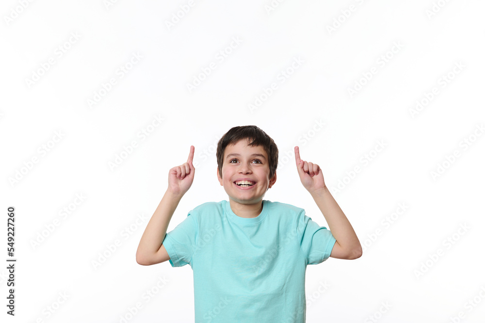 Portrait of Caucasian handsome pleasant pre-adolescent boy in blue casual t-shirt, smiling pleasantly, looking up, pointing finger at copy space for your advertising text, isolated on white background Portrait of Caucasian handsome pleasant pre-adolescent boy in blue casual t-shirt, smiling pleasantly, looking up, pointing finger at copy space for your advertising text, isolated on white background