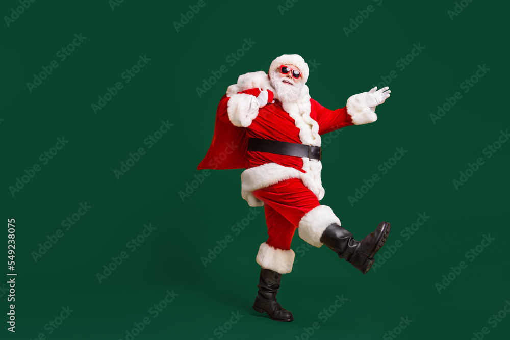 Full body profile side photo of santa claus with big belly beard carry x-mas present gift sack go wear headwear cap sunglass isolated over bright shine color background Full body profile side photo of santa claus with big belly beard carry x-mas present gift sack go wear headwear cap sunglass isolated over bright shine color background