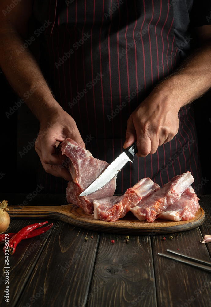 The cook cuts raw ribs with a knife on a kitchen cutting board. Barbecue grill. Idea for a restaurant or hotel menu on a dark background