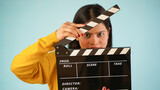 Closeup of a beautiful young Asian Indian woman standing holding clapperboard, clapper board used in film making, isolated on blue colour background studio portrait