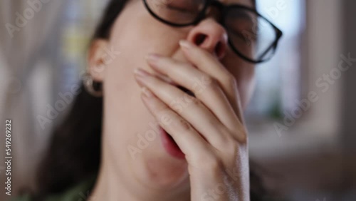 Pretty young Caucasian sleepy woman with glasses is working at laptop and yawning in close-up. Zoom in and out. Concept of overwork and boredom.