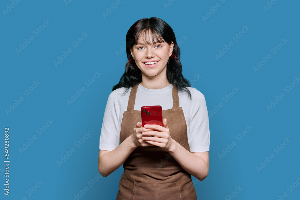 Smiling young female staff in apron using smartphone, on blue background Smiling young female staff in apron using smartphone, on blue background