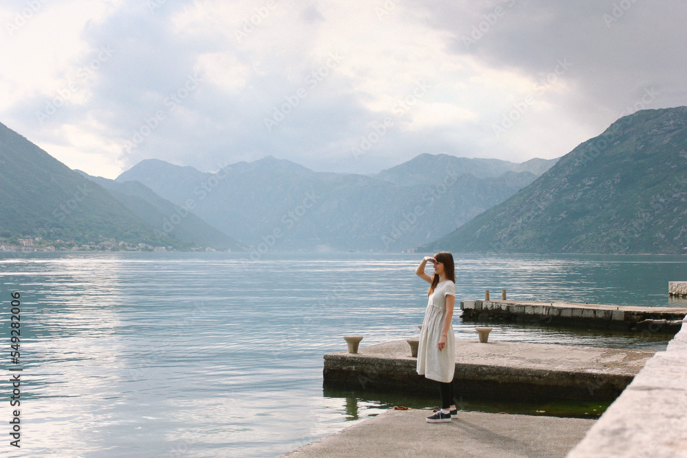 a brunette girl stands alone on the shore and looks into the distance, raising her hand to her face, peering into the sea water or the ocean against the backdrop of a mountain in montenegro