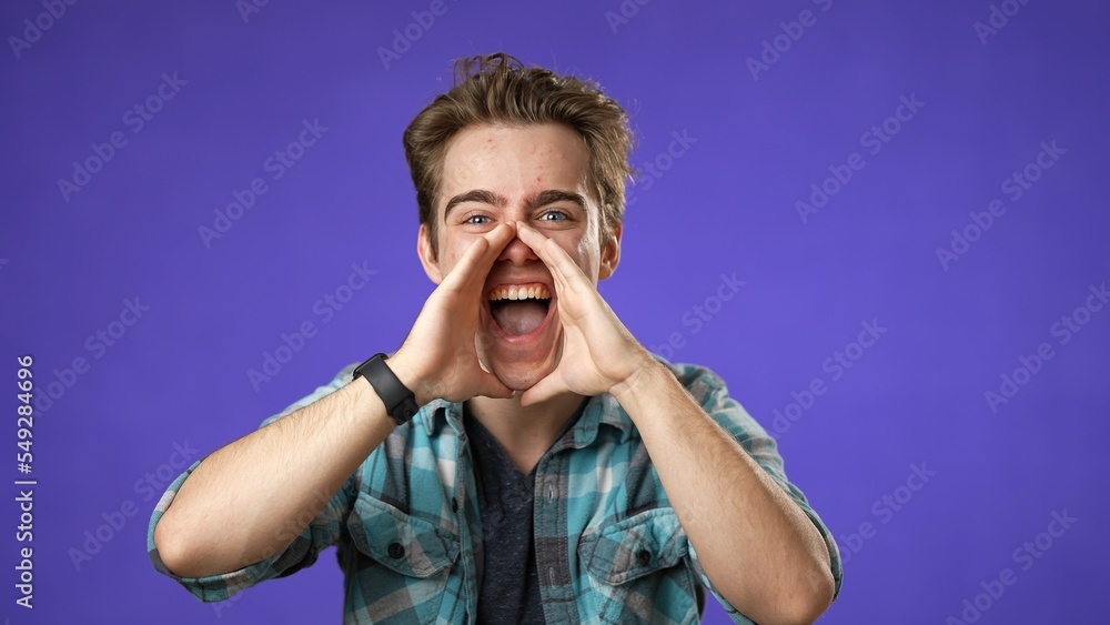 Portrait of happy young man 20s smiling scream and shout calling inviting with hands at mouth say hey you isolated on solid purple background.