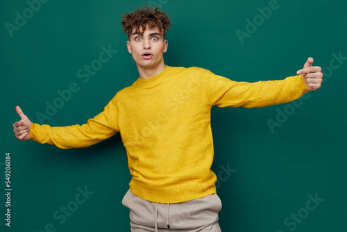 a joyful, smiling man stands on a green background in a yellow sweater and spreads his arms wide apart, making a funny face, showing thumbs up