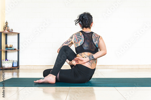 Back woman with tattoos in ardha matsyendrasana yoga pose in front of a white wall with yoga elements photo