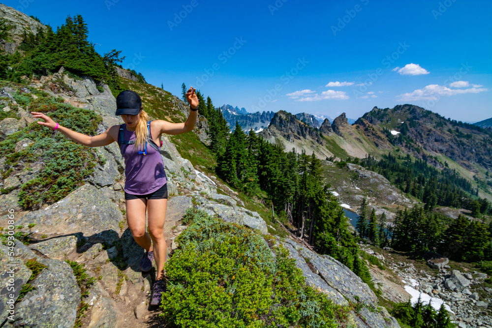 Obraz premium Adventurous athletic woman hiking down a mountain, with a beautiful mountain range in the background on a beautiful sunny day in the Pacific Northwest.