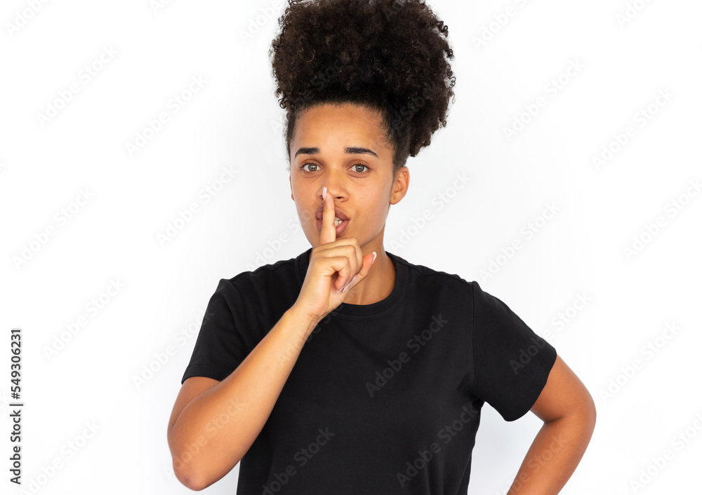 Portrait of confident young woman posing with finger on lips against white background. African American woman wearing black T-shirt making silence gesture. Secret and silence concept Portrait of confident young woman posing with finger on lips against white background. African American woman wearing black T-shirt making silence gesture. Secret and silence concept