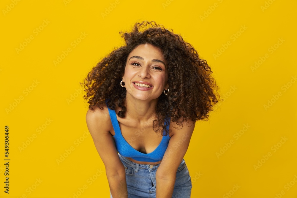 Woman with curly afro hair in a blue t-shirt on. yellow background signs with her hands, look into the camera, smile with teeth and happiness, copy space Woman with curly afro hair in a blue t-shirt on. yellow background signs with her hands, look into the camera, smile with teeth and happiness, copy space