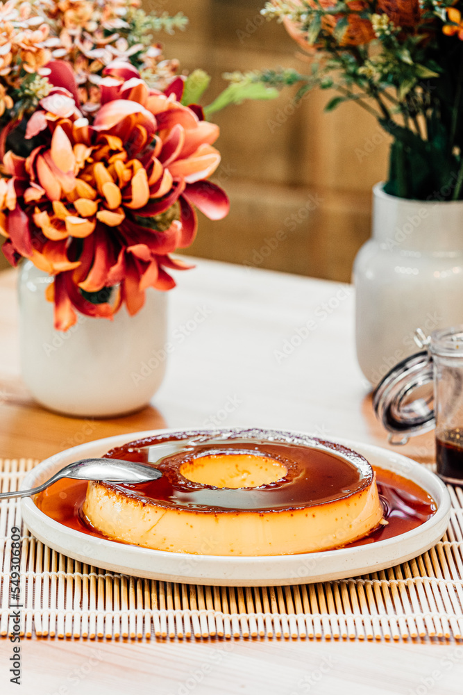 Caramel flan pudding on a plate with a spoon, flowers and plants and a cup of caramel Caramel flan pudding on a plate with a spoon, flowers and plants and a cup of caramel