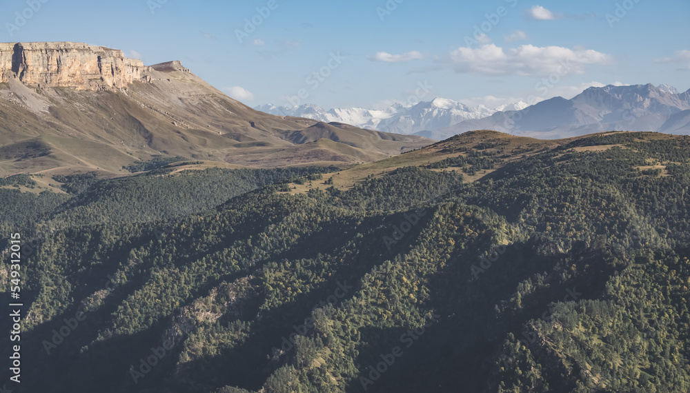 Panorama in the evening in the mountains of the Caucasus on a steep rocky slope of a table mountain against the backdrop of a mountain range with snow, hills with vegetation below