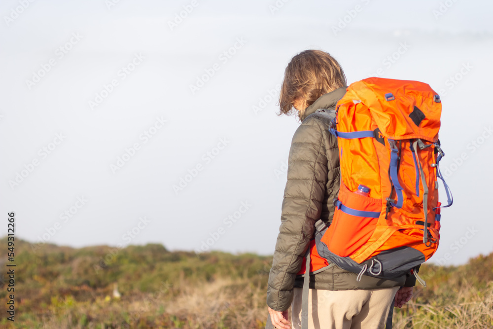 Back view of female hiker on summer day. Woman with big orange backpack standing at meadow. Hobby, active lifestyle concept