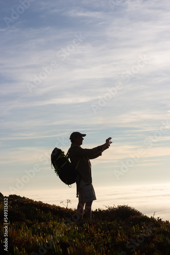 Silhouetted hiker alone at sunset. Man with backpack taking pictures with mobile phone. Hobby, nature, leisure, technology concept © KAMPUS