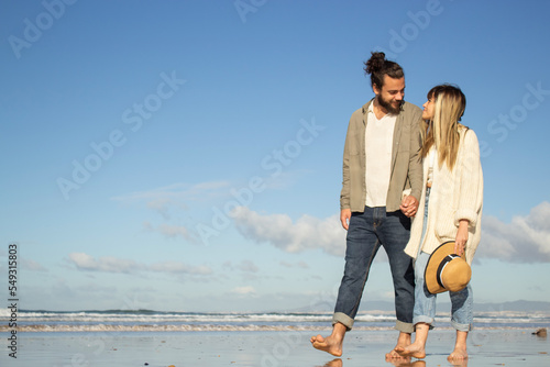 Smiling couple walking near water in summer. Bearded man and woman with hat looking at each other, holding hands. Love, travelling, dating concept