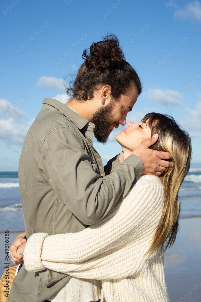 Obraz premium Romantic couple spending time at seashore on sunny day. Tall guy with man bun holding girls face in his hands, asking for kiss while happy young woman hugging her boyfriend. Romance, leisure concept