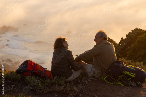 Experienced hikers at sunset. Man and woman in casual clothes and with ammunition sitting at peak, looking at each other. Hobby, active lifestyle, love concept