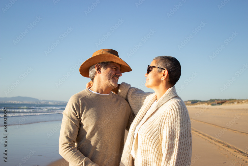 Lovely senior couple standing at seashore, hugging and looking at each other. Man in cowboy hat and short-haired woman in sunglasses enjoying vacation at seaside together. Leisure, retirement concept