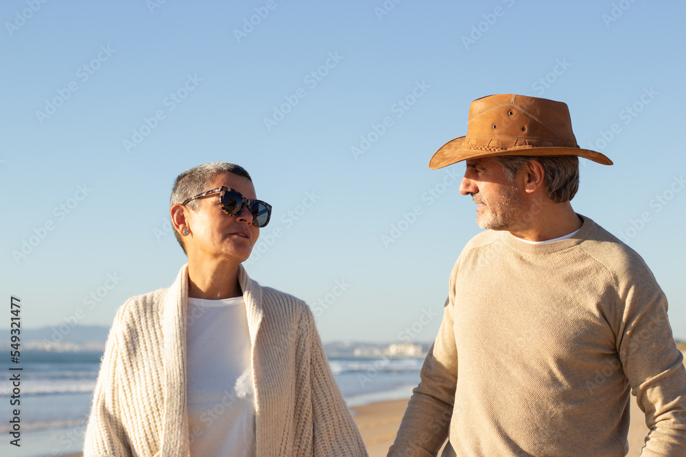 Lovely senior couple walking along seashore while enjoying holiday on sunny day. Man in cowboy hat and short-haired lady in sunglasses talking and looking at each other. Retirement, holiday concept