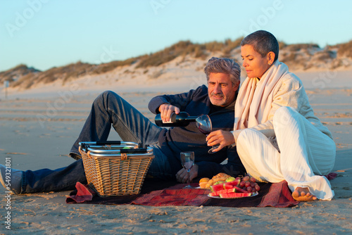 Romantic senior couple enjoying picnic with fruit and wine at seashore in the evening, sitting on blanket. Handsome grey-haired man pouring red wine into womans glass. Romance, love concept
