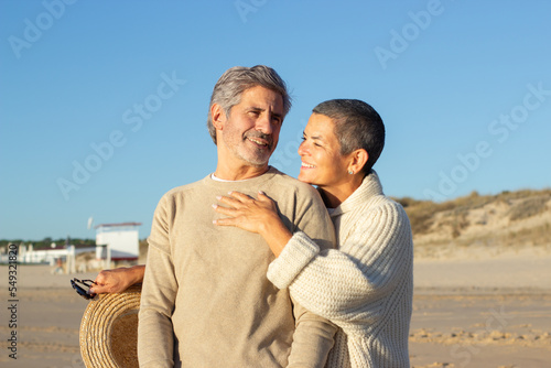 Joyful senior couple enjoying time together at the beach. Short-haired smiling woman standing behind grey-haired man and hugging him, showing affection. Relationship, love concept