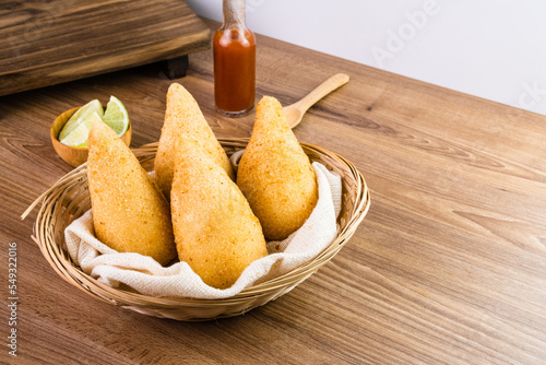 Chicken Coxinha, known as Coxinha in Brazil. Served in the basket, with spices such as lemon and pepper on the side. Wooden table in the background. Traditional brazilian snack Selective focus