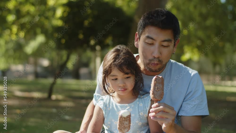 Happy Asian father and daughter eating choc ice in park. Young man sitting with little child outdoors eating ice cream. Fatherhood and childhood concept Happy Asian father and daughter eating choc ice in park. Young man sitting with little child outdoors eating ice cream. Fatherhood and childhood concept